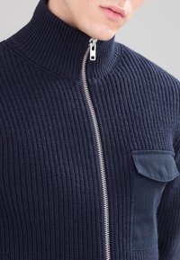 Granatowy sweter z prążkowanego dzianiny zapinany na zamek, z wysokim kołnierzem, ozdobiony srebrnym zamkiem błyskawicznym i naszywaną kieszenią na piersi. Tkanina o fakturze i solidna konstrukcja.