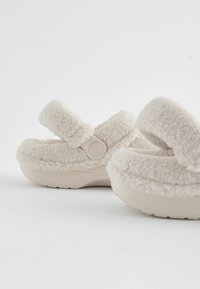 Chaussons doux, beiges, avec une texture duveteuse, une sangle ajustable et une semelle ronde et rembourrée. Convient à un usage intérieur.