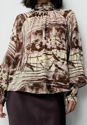 Femme portant un chemisier à col montant et manches longues avec des motifs abstraits marron et beige et une jupe sombre.