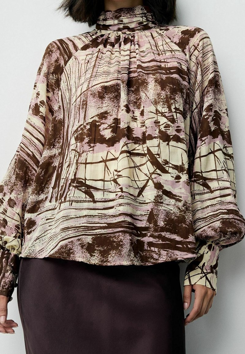 Femme portant un chemisier à col montant et manches longues avec des motifs abstraits marron et beige et une jupe sombre.