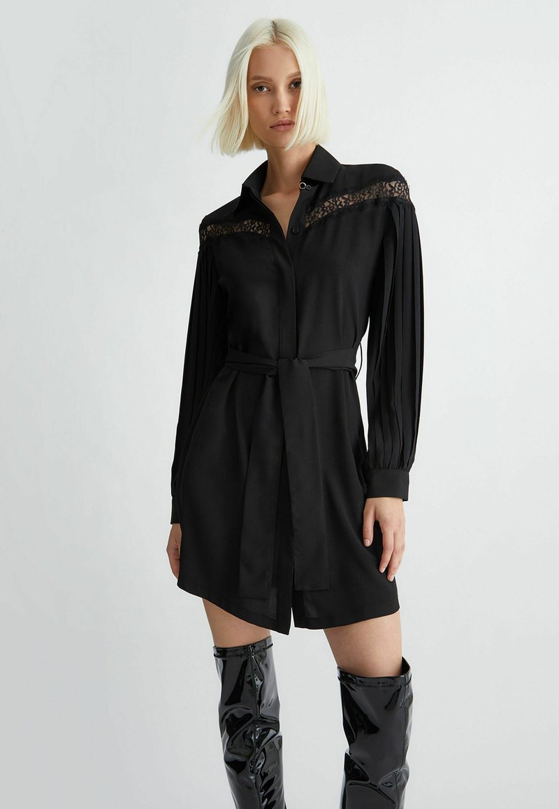 Camicia nera con dettagli in pizzo sulle spalle, maniche lunghe a pieghe, vita cinchata con cintura in tessuto e orlo dritto.