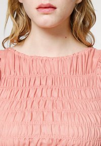 Persikkavärinen smokkikuvioinen toppi, jossa on kerättyä tekstuuria, pyöreä kaula-aukko ja lyhyet kerätyt hihat. Puuvillasekoitekangasta, kevyt ja joustava.