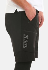 Pantaloni corti neri con una tasca con zip, tessuto testurizzato e un dettaglio logo bianco. Presentano una vita aderente e un fitting morbido.