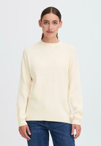 Cremefarbene Strickjacke mit hochgeschlossenem Kragen, langen Ärmeln und Rippstruktur; kombiniert mit dunkelblauen Jeans.