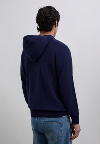 Man standing with back to camera wearing a navy blue hooded sweater and blue jeans against a plain light gray background.