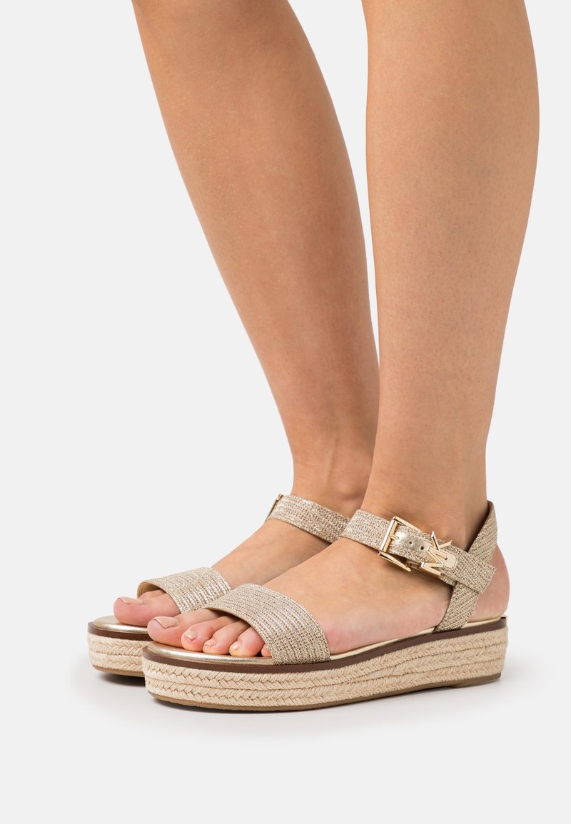 Pieds portant des sandales compensées beige tissées avec des brides à la cheville et une boucle dorée sur un fond clair.