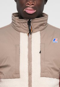 Tan and cream jacket with a high collar, textured fleece front, zip closure, and patch logo on the upper left chest.