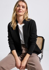 Černá strukturovaná bunda s mandarin límcem, zapínáním na knoflíky, nošena přes bílou košili. Model sedící, oblečený ve světle hnědých kalhotách.