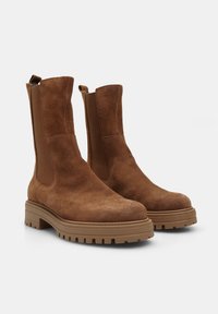 Botine din suede maro până la gleznă, cu panouri elastice pe laterale, vârfuri rotunjite și tălpi groase cu crampoane pentru aderență. Include bucle de tragere pentru a facilita încălțarea.