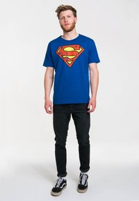 Modrá bavlněná tričko s velkým retro červeným a žlutým logem Supermana na přední straně. Model má na sobě černé džíny a tenisky.