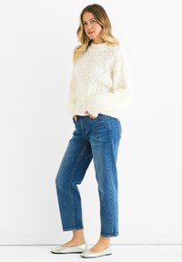 Maglione lavorato a maglia crema con collo alto, grandi motivi testurizzati, abbinato a jeans blu a gamba dritta e ballerine argentate.