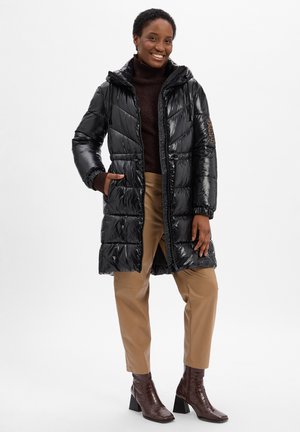Zwarte pufferjas met een glanzende afwerking, capuchon en ritssluiting. Voorzien van elastische boorden en zijzakken. G gedragen met beige broek en bruine haklaarzen.