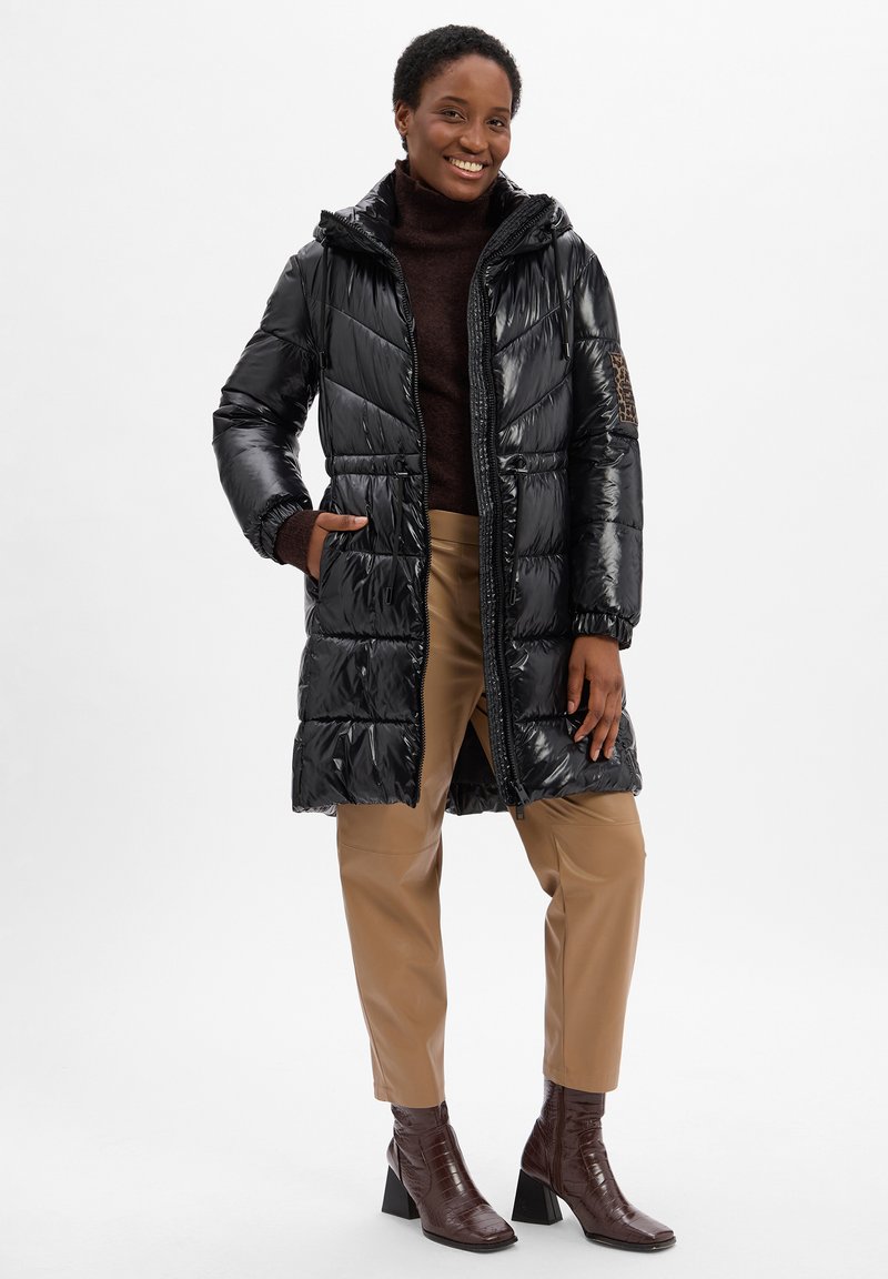 Schwarze Daunenjacke mit glänzendem Finish, Kapuze und Reißverschluss. Mit elastischen Bündchen und seitlichen Taschen. Getragen mit beigen Hosen und braunen Absatzstiefeln.