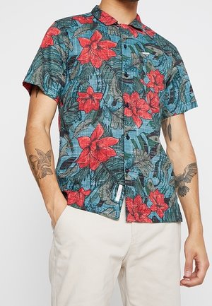 Kurzärmeliges Hemd mit Knöpfen, großen roten Blumen und grünen Blättern, getragen von einer tätowierten Person mit Händen in beigen Hosentaschen.