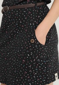 Svart klänning med ett fläckigt mönster av små rosa, vita och teal-former. Har en sidficka och ett brunt flätat bälte med en dekorativ knapp.