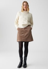 Maglione lavorato a maglia spesso con colletto alto, abbinato a una gonna in velluto a coste marrone con tasche frontali e stivaletti neri.
