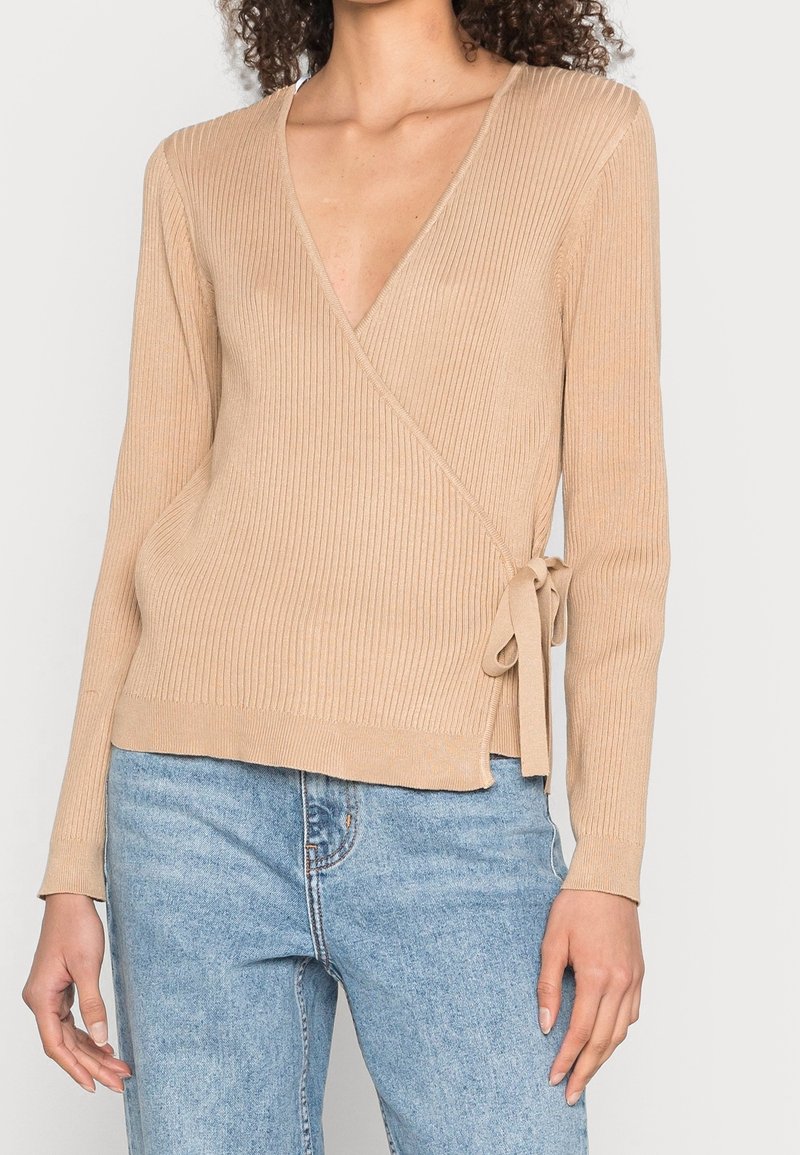 Femme portant un pull beige côtelé à nouer sur le côté et un jean taille haute bleu clair, debout devant un fond blanc.
