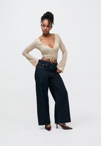 Bēša garroku wrap stila topiņa ar auklas akcentu, kombinēta ar tumši zilām plašām džinsa biksēm un smailiem bordo papēžiem.