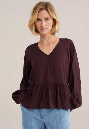 Lyshåret kvinde iført en mørk lilla langærmet V-hals bluse med indsnøret talje og lyseblå jeans, stående mod en ensfarvet baggrund.