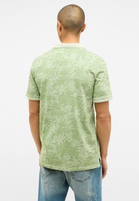 Grünes Polohemd mit einem strukturierten Blattmuster, weißem Kragen und Manschetten sowie kurzen Ärmeln. Kombiniert mit hellblauen, ausgebleichten Jeans.