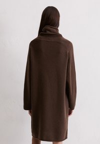 Braunes, übergro�ßes Strickkleid mit hohem Kragen, langen Ärmeln und einer weichen Textur. Der Saum ist leicht geschwungen.