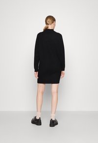 Robe noire en tricot avec des manches longues et des poignets à côtes, présentant une encolure ronde et une coupe décontractée, portée avec des chaussures sombres et des chaussettes claires.