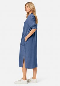Středně dlouhé modré šaty vyrobené z měkké tkaniny, s kulatým výstřihem, krátkými nařasenými rukávy, bočními kapsami a rozparkem na lemu.
