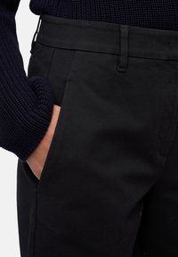 Hand in de zak van zwarte broek, gecombineerd met de linkerzijde van een donkerblauwe gebreide trui zichtbaar.