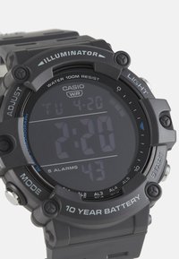 Montre numérique Casio avec un bracelet en caoutchouc noir et un boîtier rond gris. Les caractéristiques incluent un écran rétroéclairé, une résistance à l'eau et plusieurs alarmes.