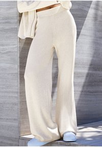 Široké nohavice s rebrovaným vzorom v svetlo béžovej pletenine. Majú vysoký pás a sú skombinované so svetlomodrými teniskami na textúrovanom pozadí.