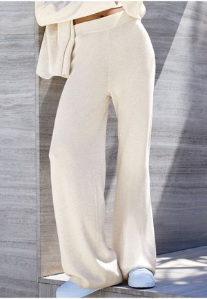 Weite, gerippte Hosen aus hellbeigem Strickstoff. Sie haben einen hohen Bund und werden mit hellblauen Sneakers auf einem strukturierten Hintergrund kombiniert.