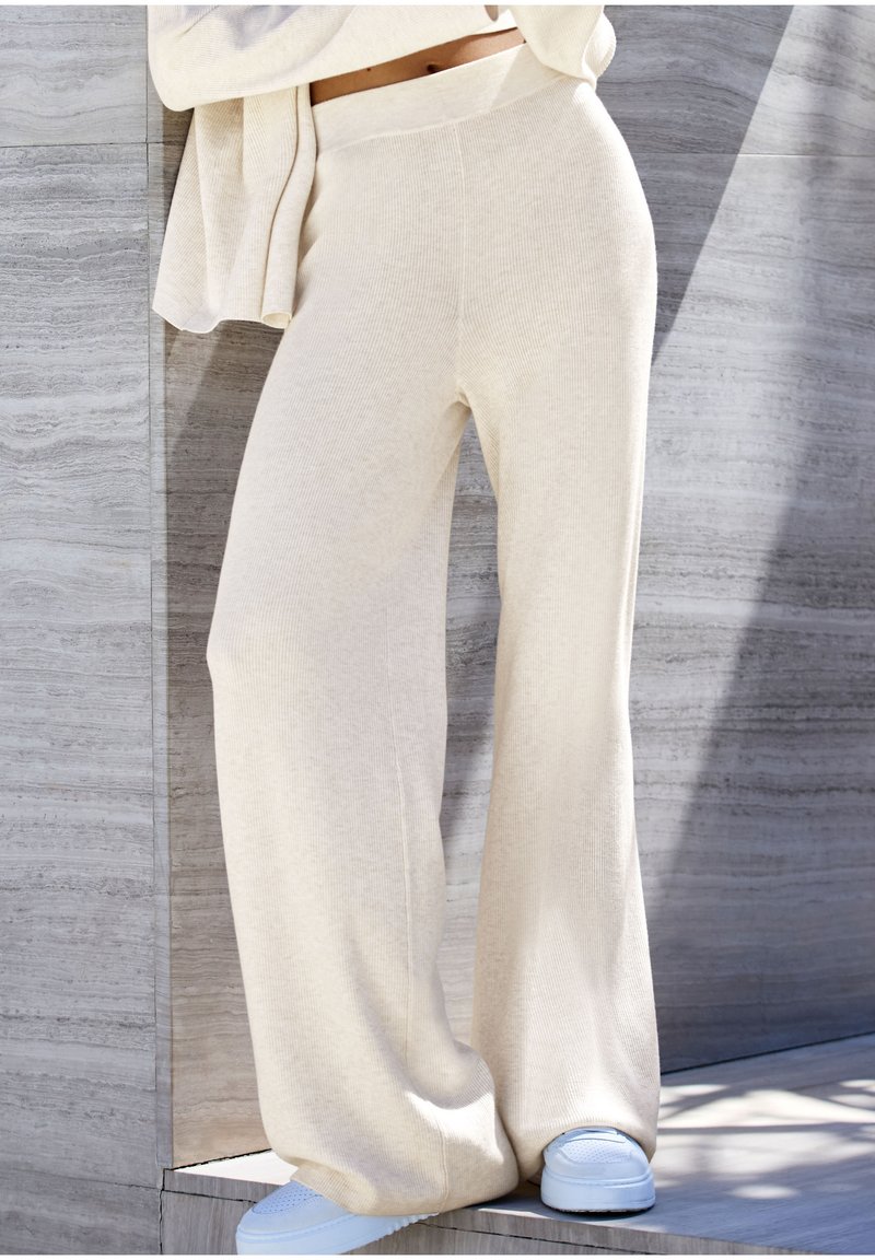 Široké nohavice s rebrovaným vzorom v svetlo béžovej pletenine. Majú vysoký pás a sú skombinované so svetlomodrými teniskami na textúrovanom pozadí.