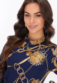 Tengerészkék ruha bonyolult aranylánc- és embléma mintákkal, sima szövet textúrával és lekerekített nyakkivágással.