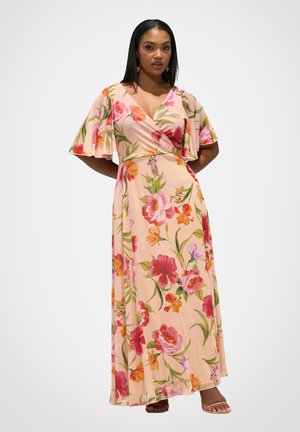Kvinde iført en lang, ferskenfarvet blomsterwrap-kjole med flæseærmer og lyserøde, hælede sandaler, stående mod en ensartet hvid baggrund.