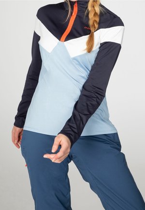 Frau mit Zopf, die ein farblich abgesetztes Langarmoberteil in Schwarz, Weiß, Hellblau und eine blaue Hose trägt und mit angewinkelten Armen vor grauem Hintergrund posiert.