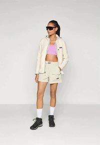 Fleece zip-jakke i off-white med mint aksenter, kombinert med en lys rosa crop topp og beige shorts. Sorte hiking-støvler fullfører looken.