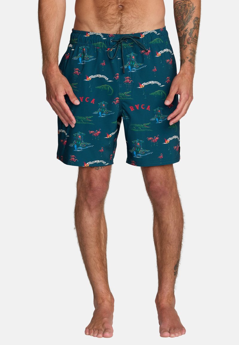 Mężczyzna stojący boso, ubrany w turkusowe kąpielówki z czerwonymi flamingami, zielonymi aligatorami, napisem "RVCA" i banerami "Welcome to Paradise".