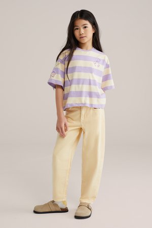 Niña joven de pie, vestida con una camiseta de rayas lavanda y crema con detalles florales, pantalones amarillo pálido y zapatos beige sin cordones.