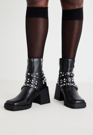 Bottes en cuir noir mi-mollet avec des clous et des sangles argentés, portées avec des bas noirs transparents s'arrêtant au genou sur des jambes à la peau foncée.