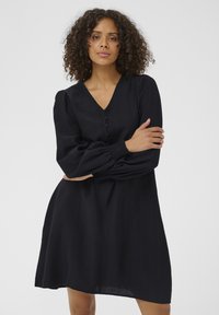 Robe noire longueur genou avec manches longues bouffantes et encolure en V. Elle présente des boutons sur le devant et une silhouette décontractée et fluide.
