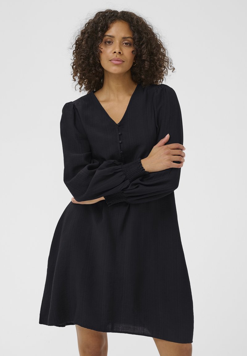 Robe noire longueur genou avec manches longues bouffantes et encolure en V. Elle présente des boutons sur le devant et une silhouette décontractée et fluide.