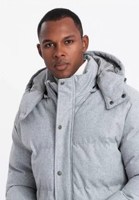 Chaqueta acolchada gris aislante con alto cuello, gran capucha y botones a presión. Textura suave con un forro liso. Presenta un diseño moderno y ajustado.