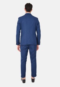 Completo blu a due pezzi realizzato in tessuto strutturato. La giacca ha un rever a punta, pantaloni slim fit con orli arrotolati, indossati con scarpe scure.