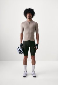Beige fietsshirt met korte mouwen, nauwsluitend ontwerp met een rits aan de voorkant. Zwarte fietsbroek, witte sokken en een witte helm. Inclusief handschoenen.