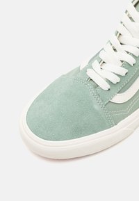 Gros plan sur une basket en daim vert clair et toile avec des lacets blancs et une semelle en caoutchouc blanche sur un fond uni.