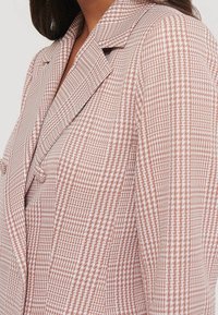 Bleiseri, jossa on vaaleanpunainen ja valkoinen houndstooth-kuvio, räätälöity istuvuus, lovet kaulukset ja kaksi etunappia. Rakenteellinen, teksturoitu kangas.