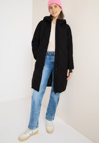Zwarte gewatteerde pufferjas met capuchon, lange mouwen en ritssluiting, gecombineerd met lichtblauwe jeans en witte sneakers met blauwe accenten.