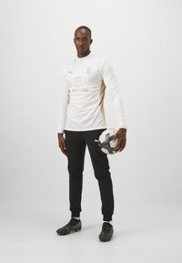 Weißes Football-Trikot mit goldenen Akzenten, langen Ärmeln und einem Viertelreißverschluss, kombiniert mit schwarzen Hosen und schwarzen Football-Schuhen, während ein Ball gehalten wird.