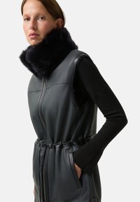 Czarna skórzana kamizelka z wysokim, puszystym czarnym futerkiem na kołnierzu, z zamkiem z przodu, taliowanym krojem i bocznymi kieszeniami; gładka tekstura, bez rękawów.