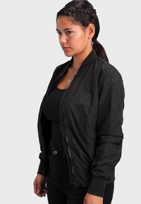 Schwarze Bomberjacke mit Reißverschluss vorne, gerippten Bündchen und einem strukturierten Patch auf der linken Brust. Kombiniert mit einem schwarzen Tanktop und hochgeschnittenen Jeans.